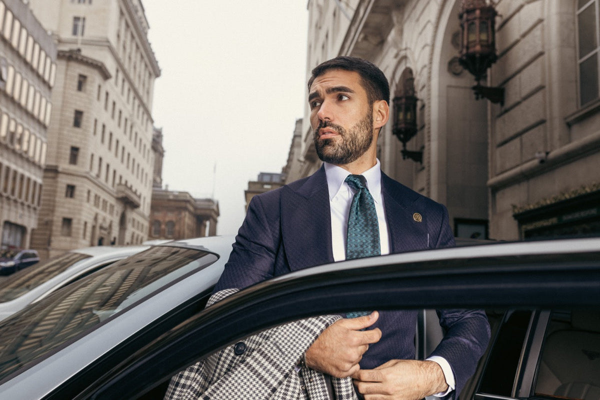 Man wearing a navy tailored suit and patterned green tie beside a car on a city street